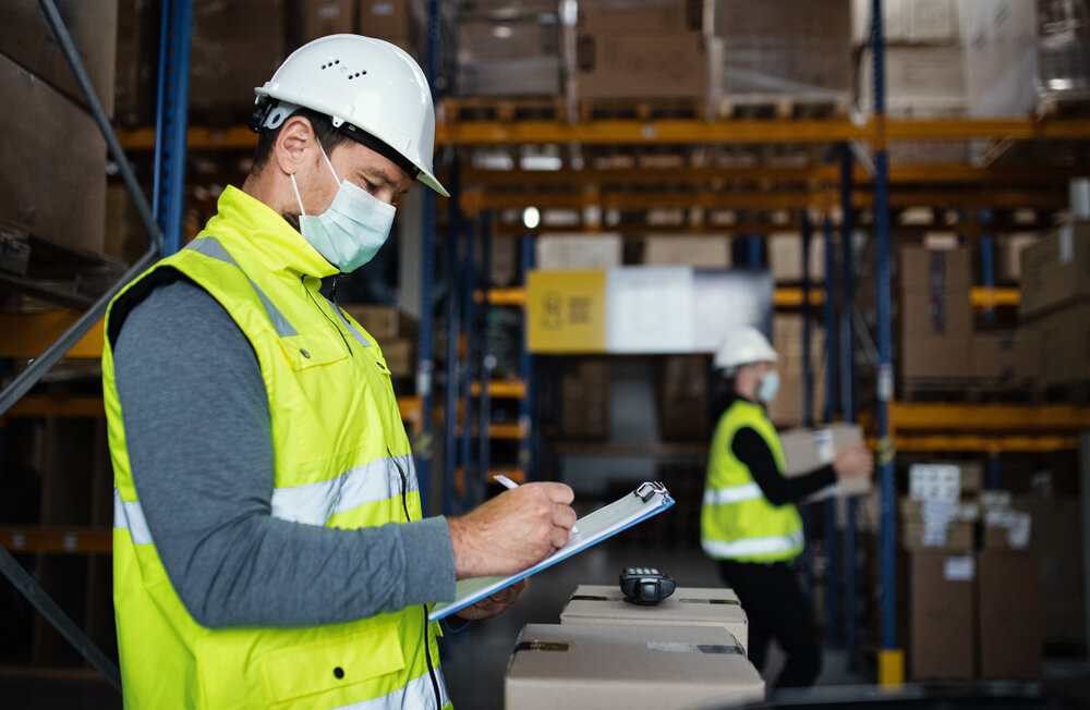 Man worker with clipboard working indoors in warehouse, wearing COVID mask